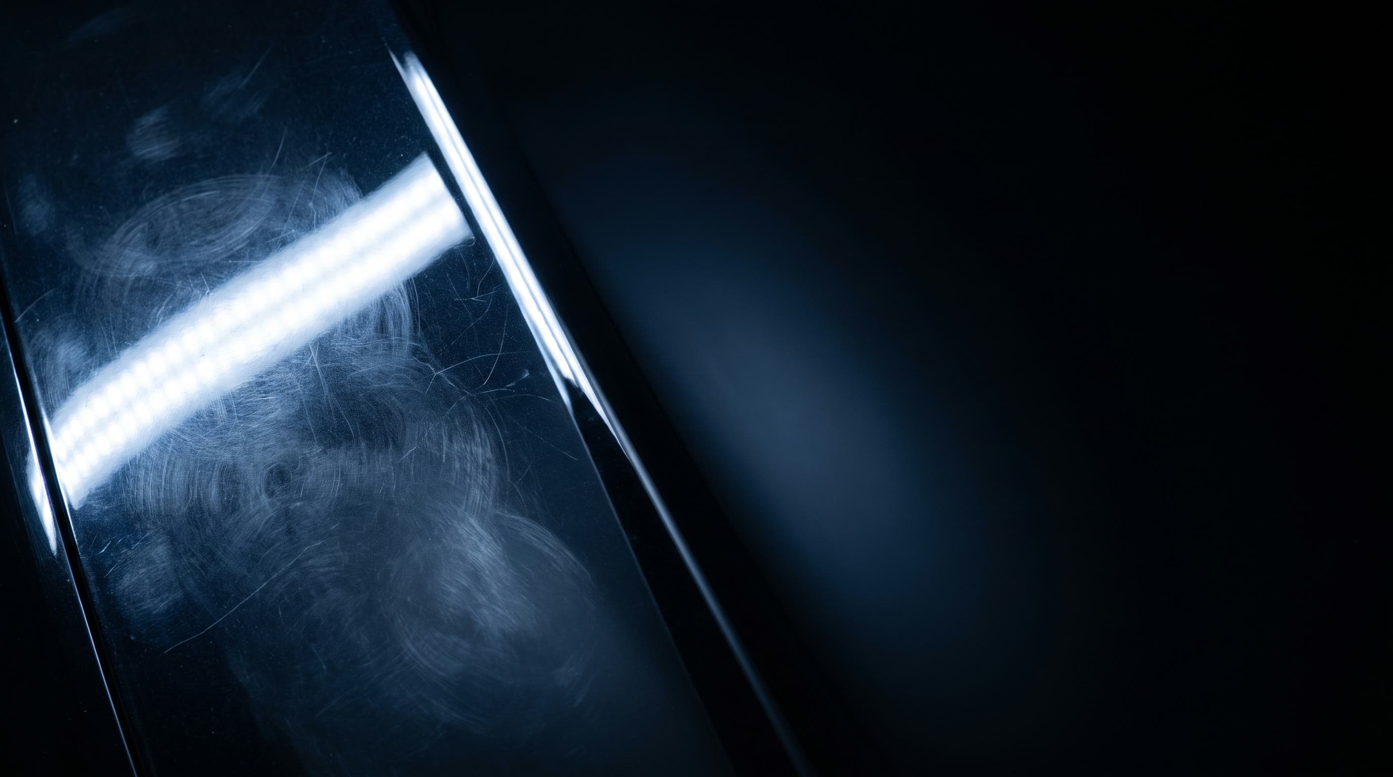 A single strip light raking across dark automotive paint, revealing swirl marks on one side and a mirror finish on the other
