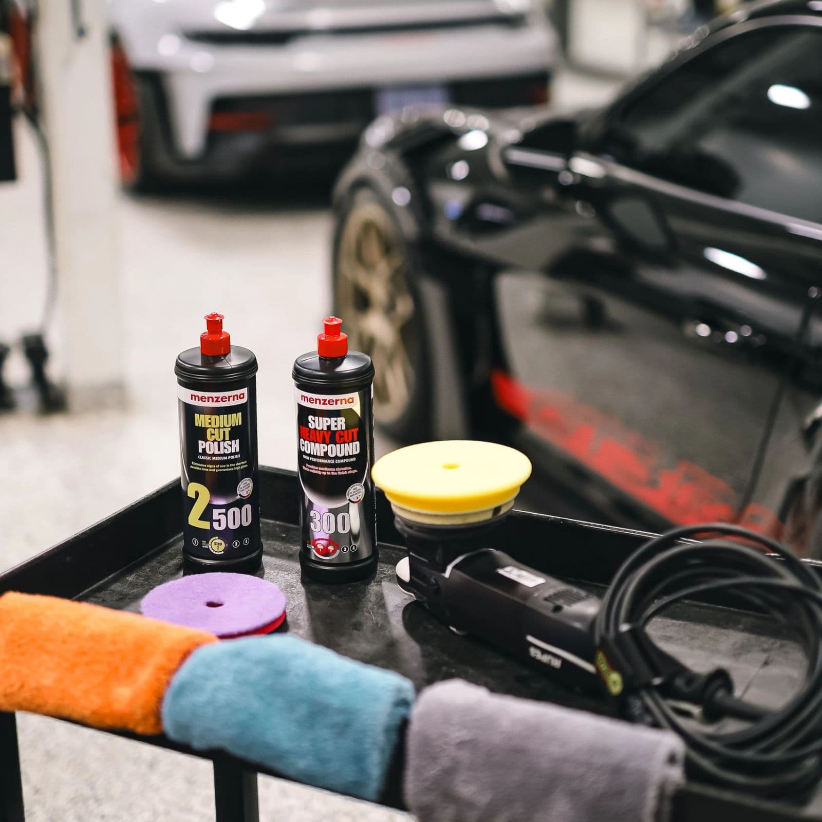 Polishing compounds, pads and a dual action polisher laid out beside a dark car